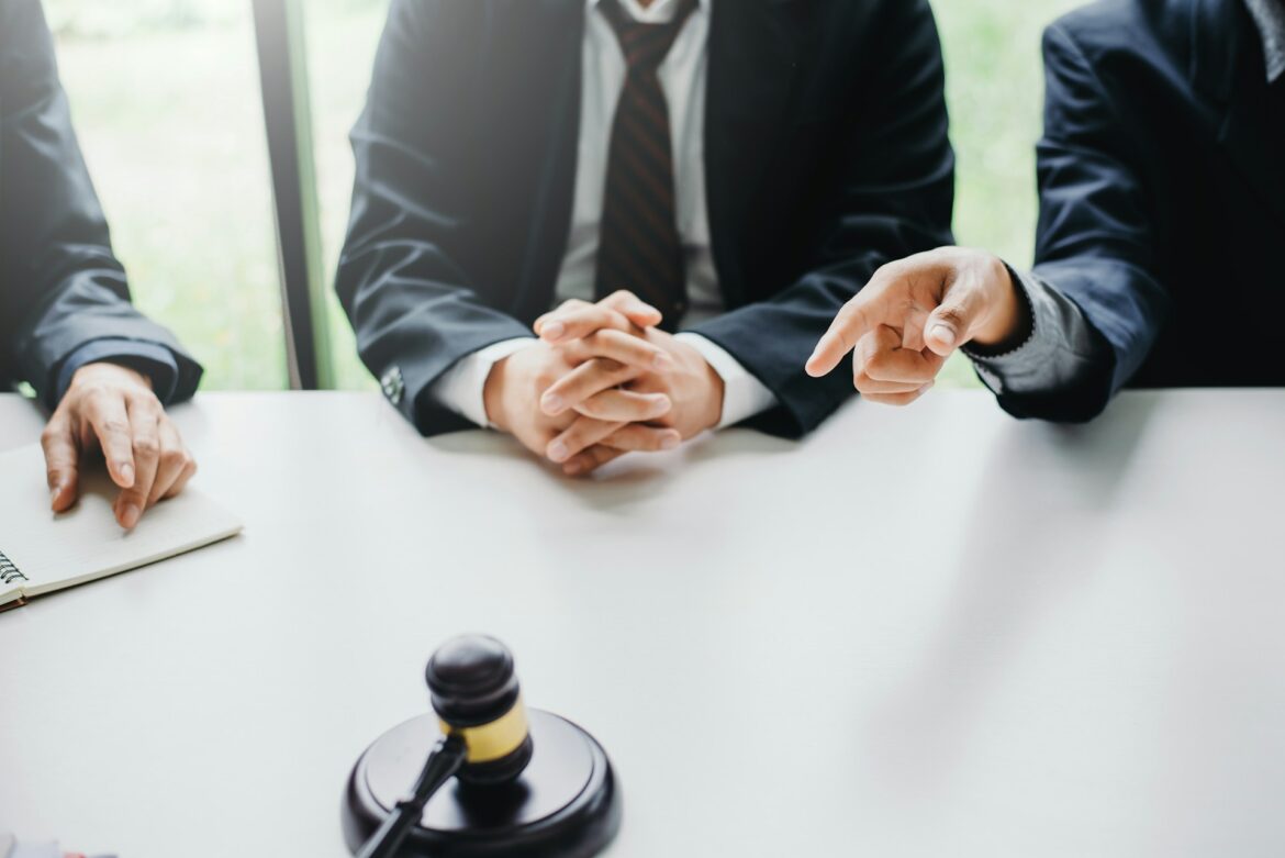 Three professionals in suits sit at a conference table with a gavel visible in the foreground, suggesting a legal meeting or proceeding.