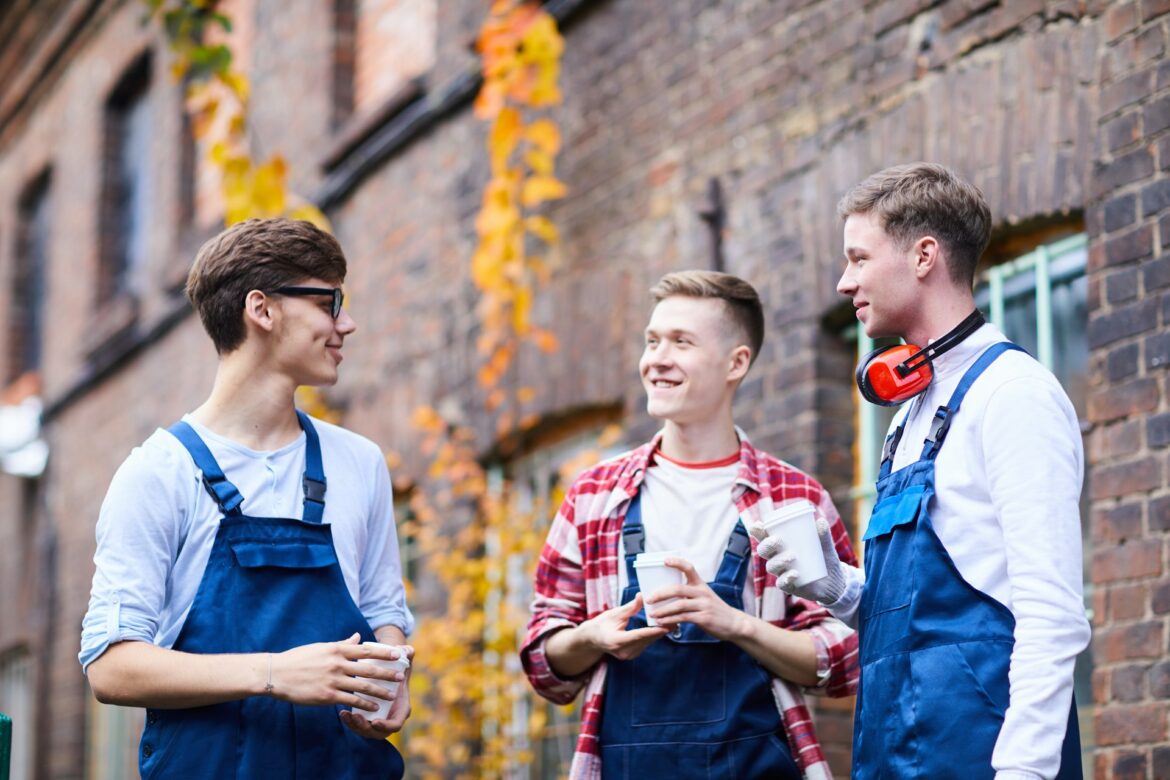 Three young men in blue overalls chat outdoors along a brick wall, holding coffee cups; autumn leaves in background.