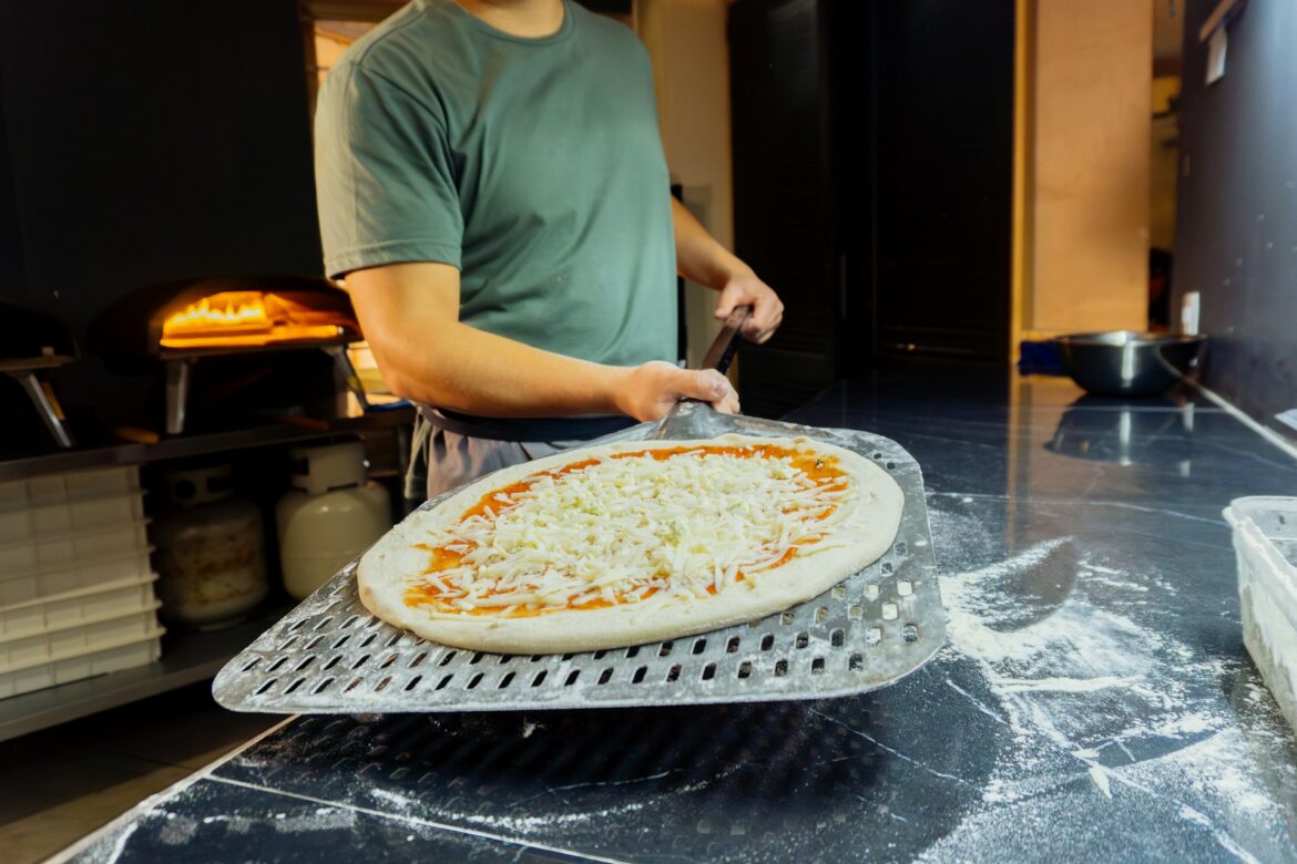 Pizza with cheese and tomato sauce on a perforated metal peel, ready to bake in a pizzeria kitchen.