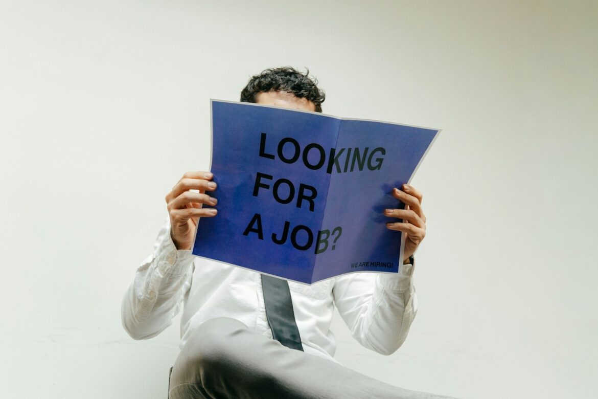 Person holding a blue flyer that reads 'LOOKING FOR A JOB?' with a small 'WE ARE HIRING' note, while sitting.