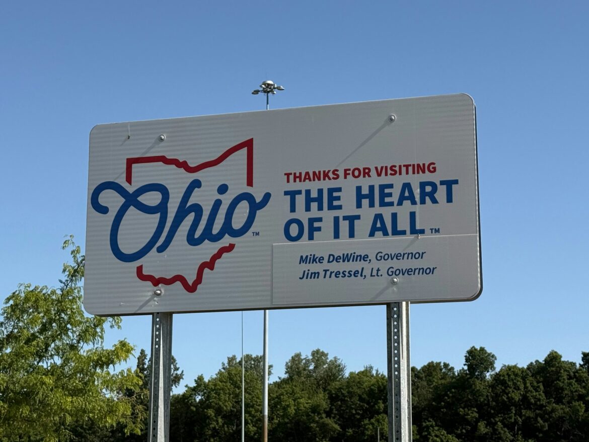 Sign reading 'Ohio' with red state outline and 'THANKS FOR VISITING THE HEART OF IT ALL' against a blue sky.