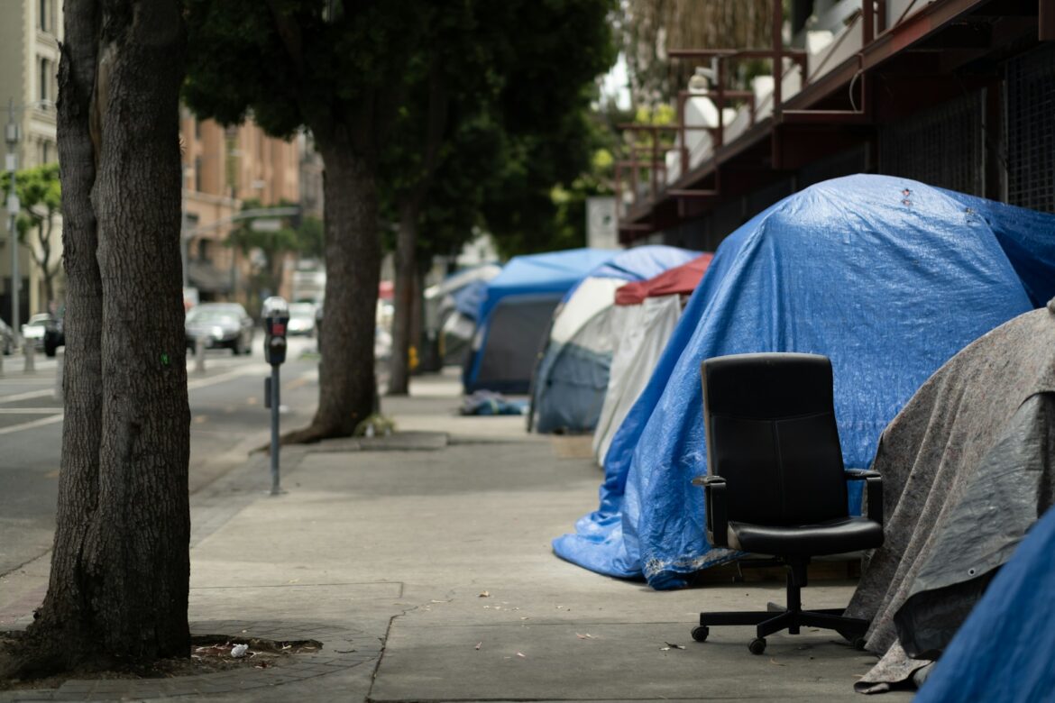 Urban sidewalk encampment with blue tarp-covered tents along a storefront and a black office chair in the foreground.