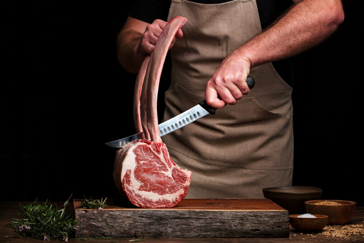Butcher carving a large rib bone from raw meat with a knife, rosemary nearby on a wooden table.