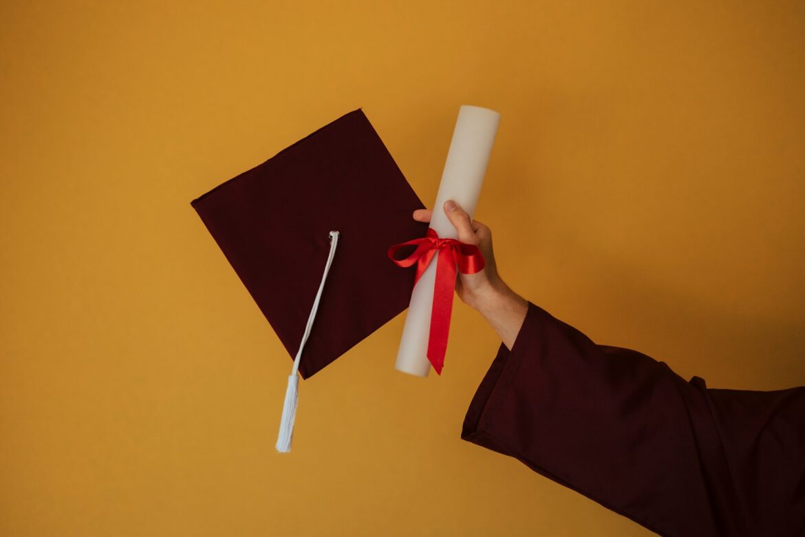 Graduate’s hand holds a rolled diploma with a red ribbon beside a maroon graduation cap against an orange backdrop