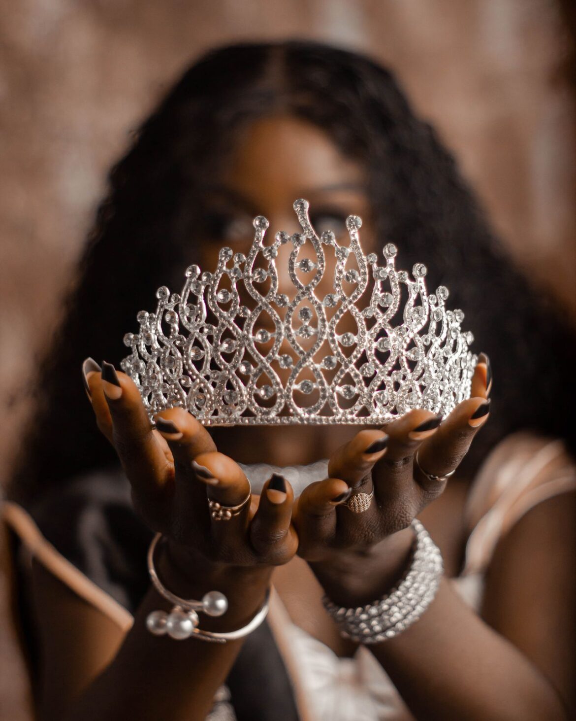 Close-up of a jeweled tiara being held up by two hands, with the person's face softly blurred in the background and wearing multiple bracelets and rings.