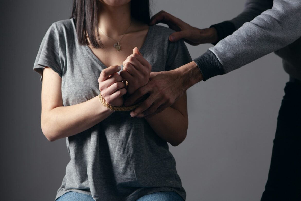Woman with her wrists bound by rope in front, a second person resting a hand on her shoulder against a gray background, tense scene.