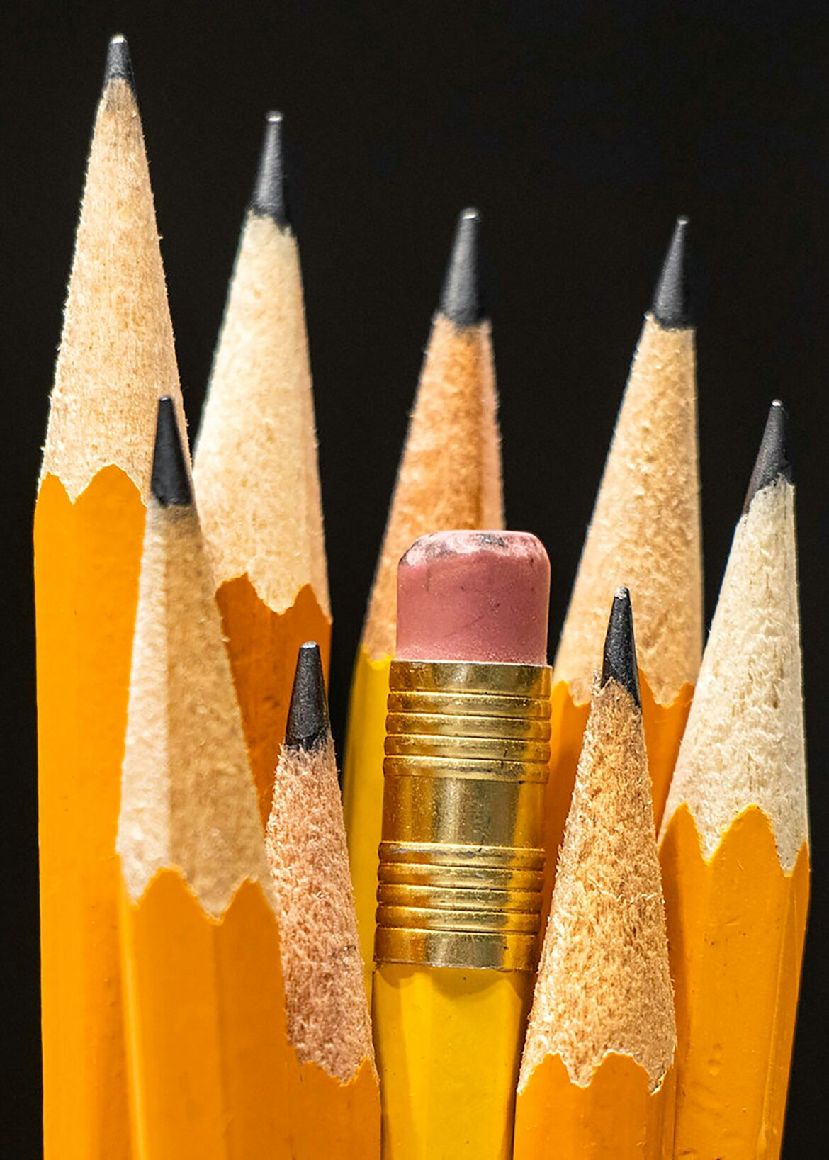 Close-up of sharpened yellow pencils with graphite tips and a pink eraser in the center against a black background.