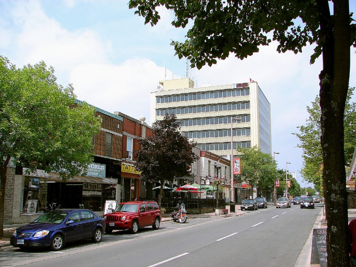 City street with small shops, parked blue sedan and red SUV, and a tall beige office building in the background.