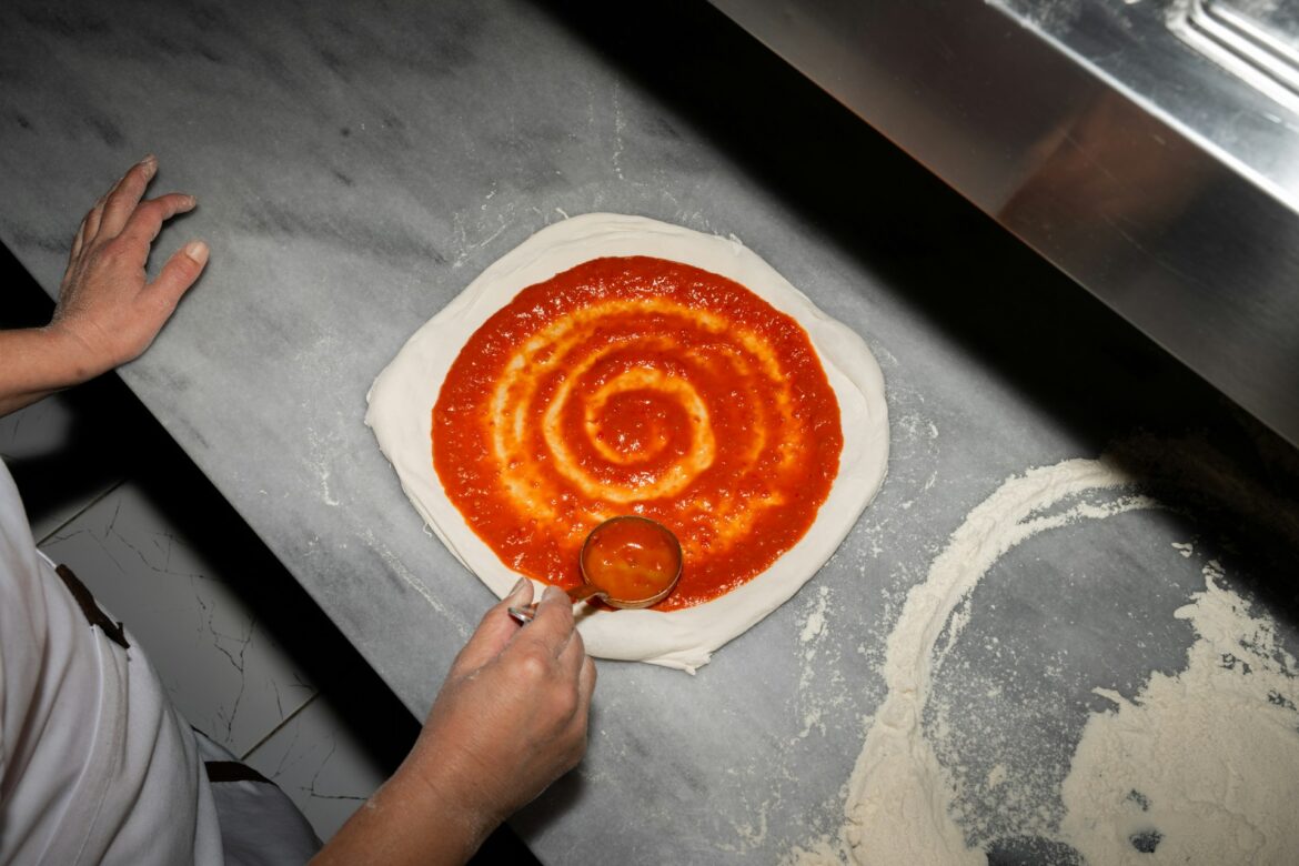 Person spreads tomato sauce over a round pizza dough on a stainless prep table, creating a spiral pattern before adding toppings.