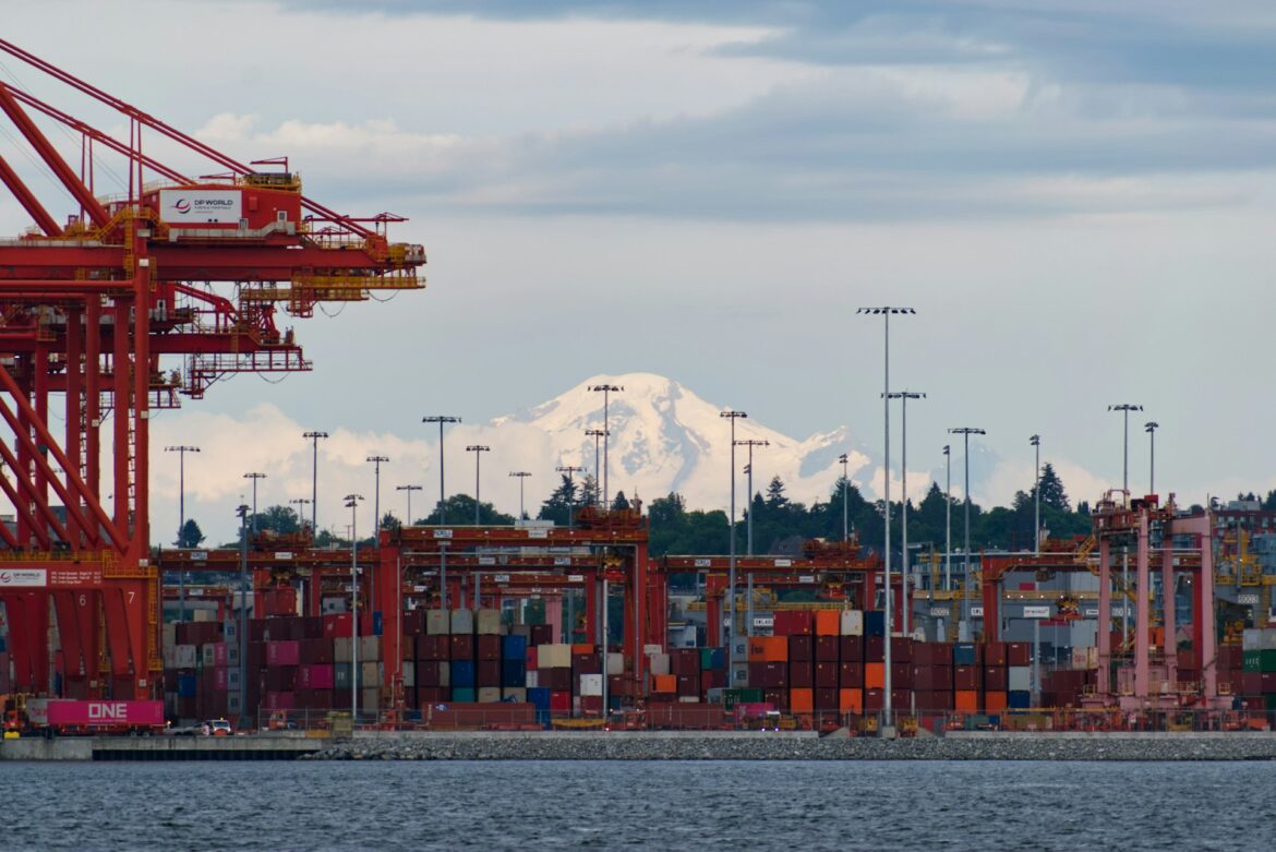 Shipping port scene with tall red gantry cranes and stacked colorful shipping containers, mountain range visible in the background.