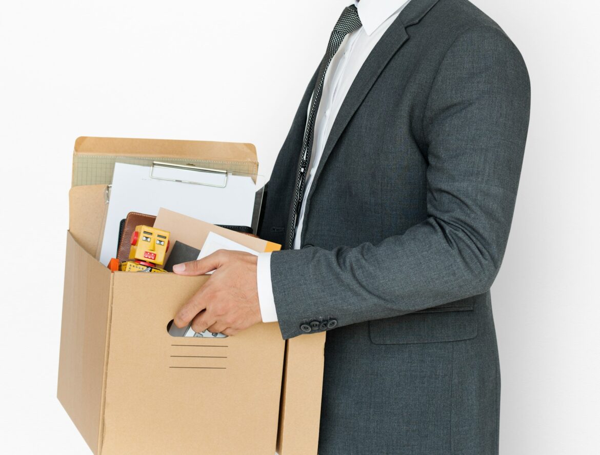 Person in a gray suit holding a cardboard box filled with papers and office supplies, including a small toy robot inside the box.
