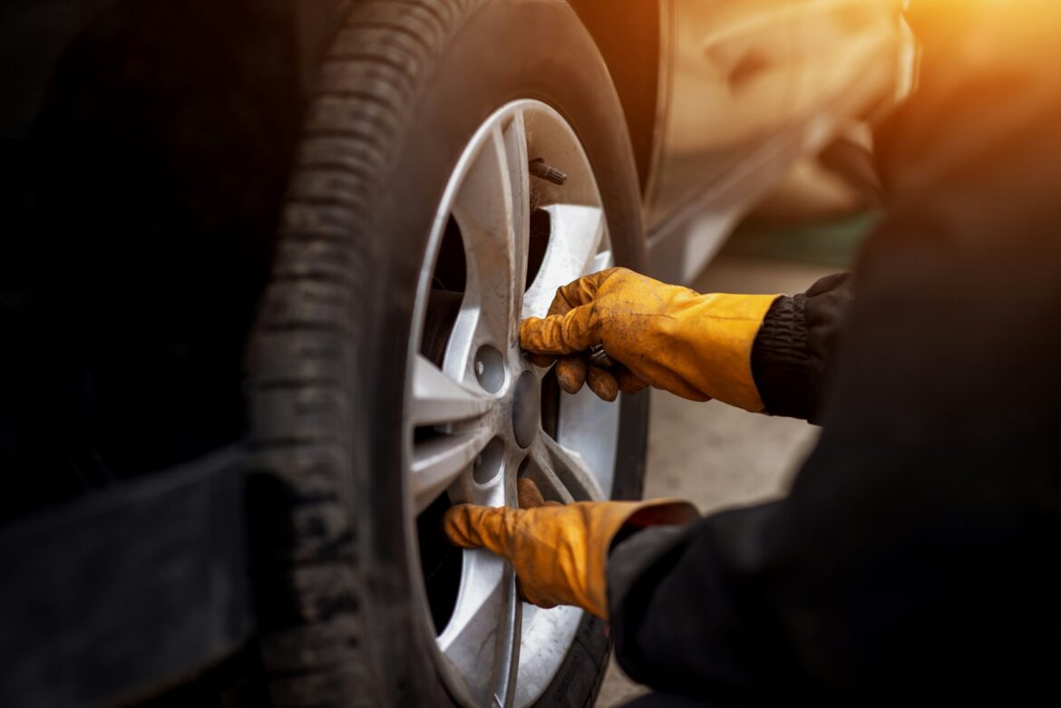 Mechanic wearing orange gloves tightening lug nuts on a car wheel outdoors as sunlight glows to the right.