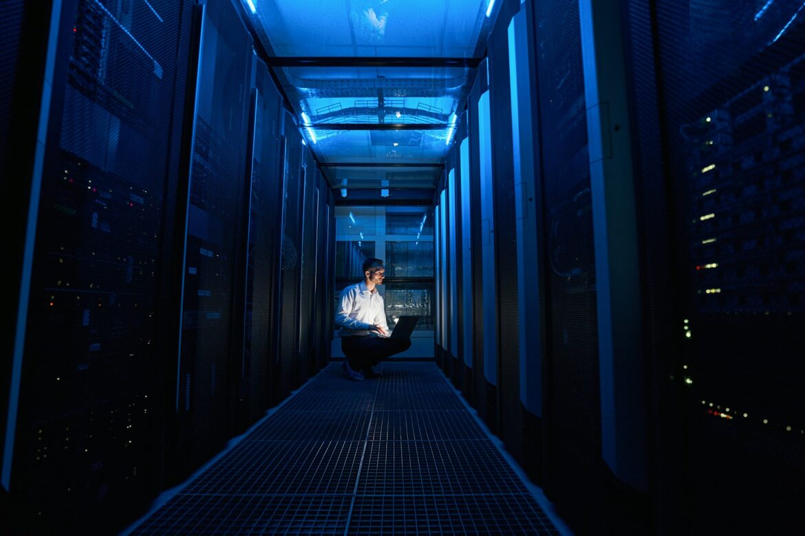 Man kneeling with a laptop in a blue-lit data center corridor between server racks.