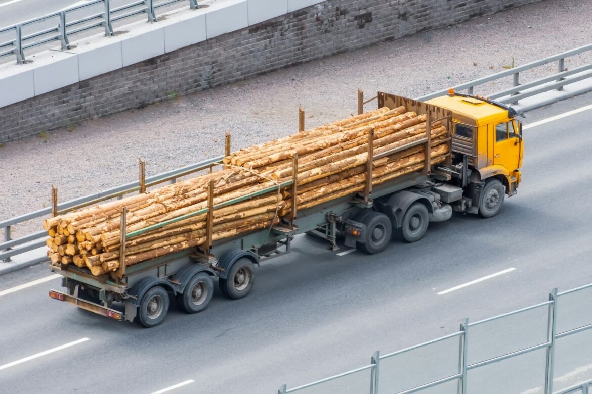 Yellow logging truck on a highway carrying long bundled logs secured on a flatbed trailer, with guardrails nearby