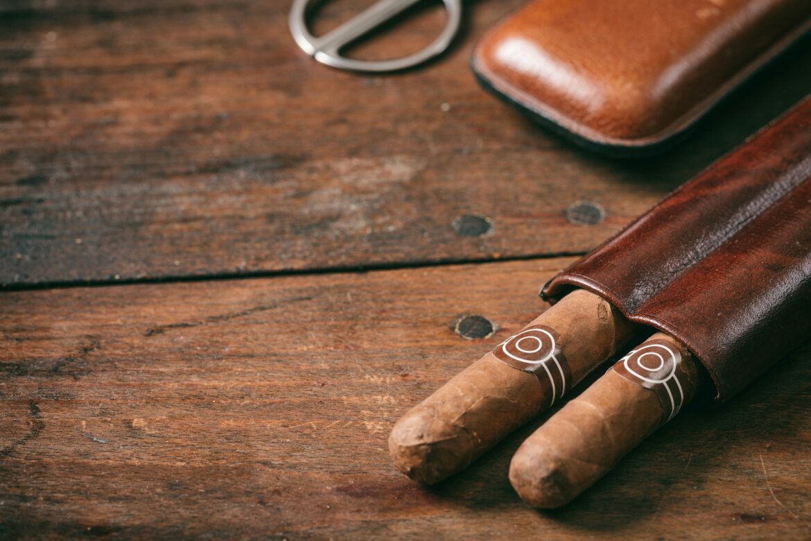 Two cigars with decorative bands rest on a weathered wooden surface beside a brown leather cigar case or roll-up sleeve edge and a metal cutter in the background.