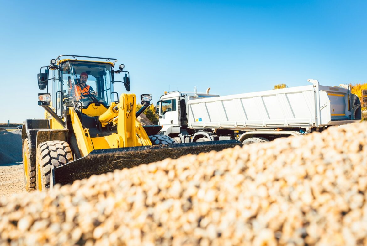 Yellow front-end loader at a construction site with a white dump truck nearby, under a clear blue sky, and a worker in the cab wearing an orange vest.