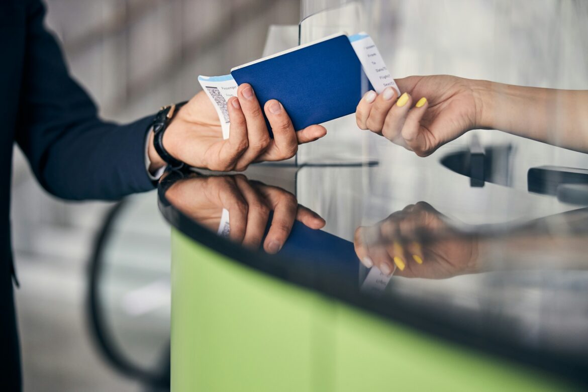 Two hands exchange a blue passport-like booklet and receipts over a glass counter at a service desk.