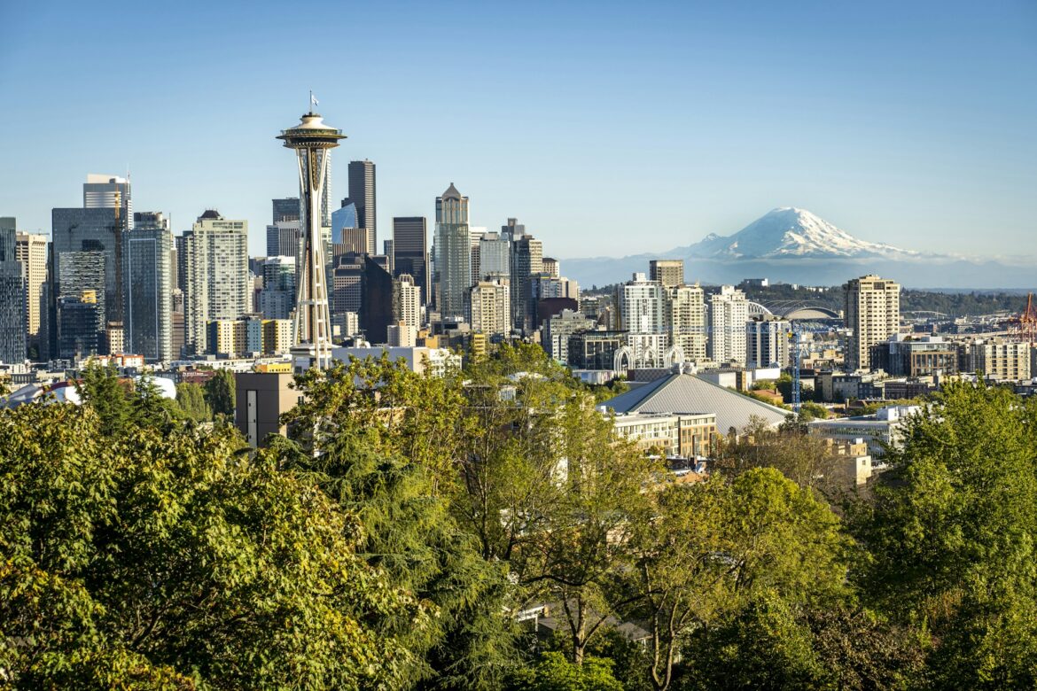 Seattle skyline with Space Needle, high-rise buildings, and Mount Rainier in the distance framed by green trees in the foreground.