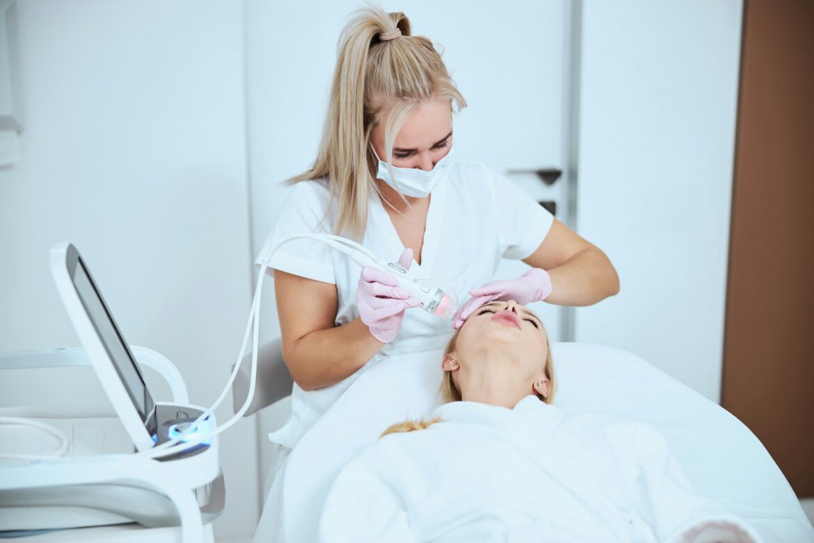 Clinician in a white uniform and mask performing a facial treatment on a patient lying in a clinic, using a handheld device near the face.