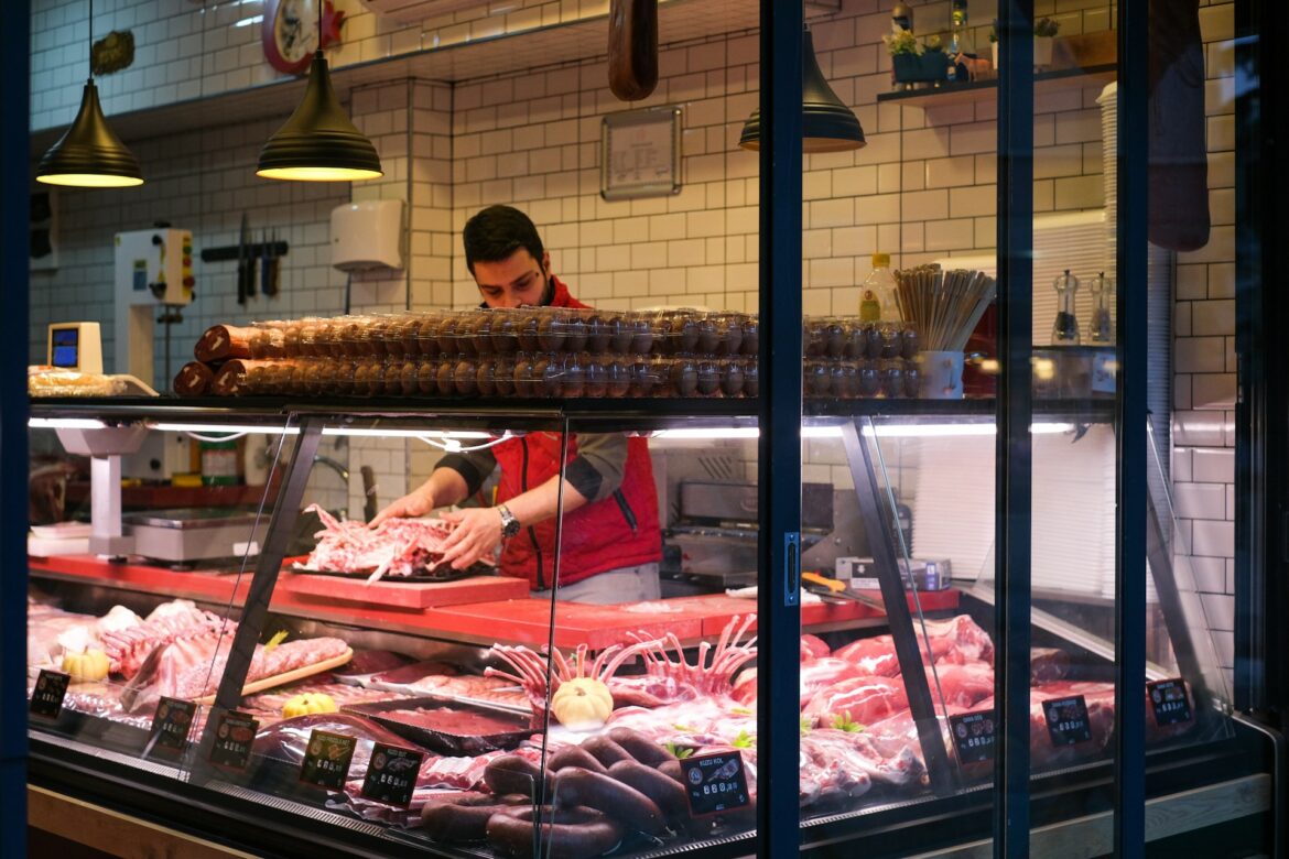 Butcher in a red vest slices meat behind a glass deli counter with sausage stacks above.