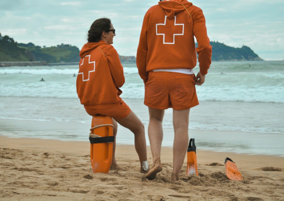 Two lifeguards in orange uniforms standing on a sandy beach, facing the sea with rescue gear nearby.