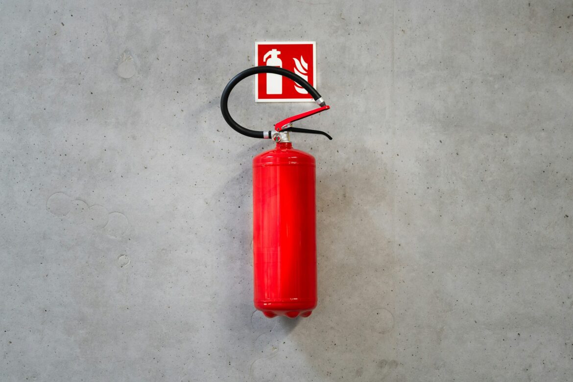Red fire extinguisher mounted on a gray concrete wall with a red fire-safety sign behind it.