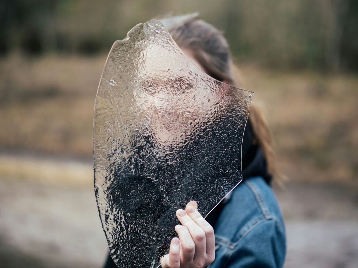 Person holding a large, jagged, reflective shard up to their face outdoors.