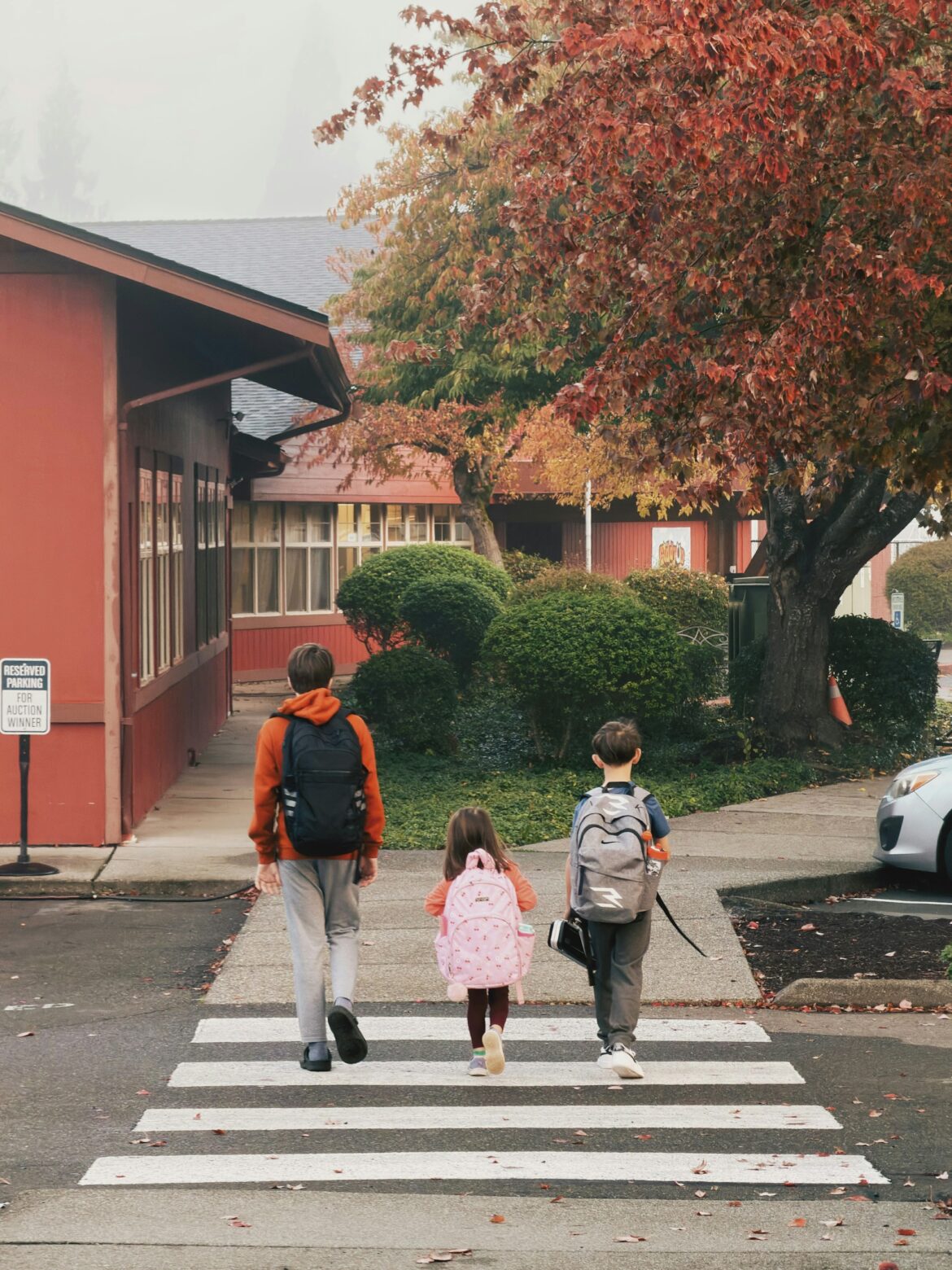 Children with backpacks walk across a zebra crosswalk toward a red building amid autumn trees with red and orange leaves.