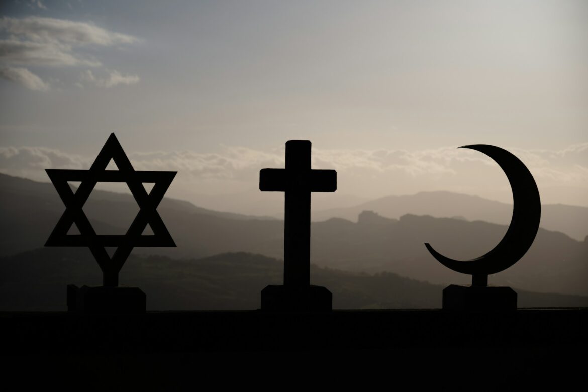 Silhouettes of a Star of David, a Christian cross, and a crescent moon against a sunset over rolling hills.