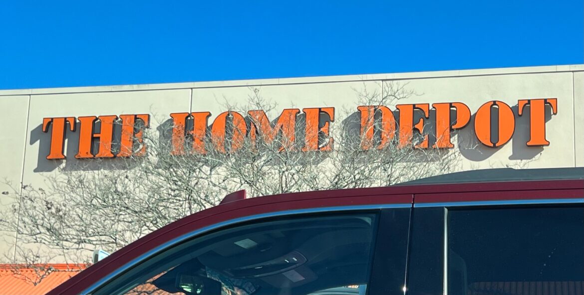 The Home Depot store sign in orange letters on a beige wall, with leafless branches in front and a red vehicle in the foreground.