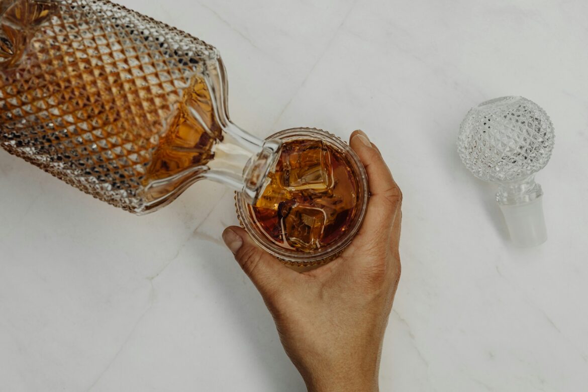 Top-down view of a hand pouring amber whiskey from an ornate crystal decanter into a glass with ice on a white marble surface; a crystal stopper sits nearby.