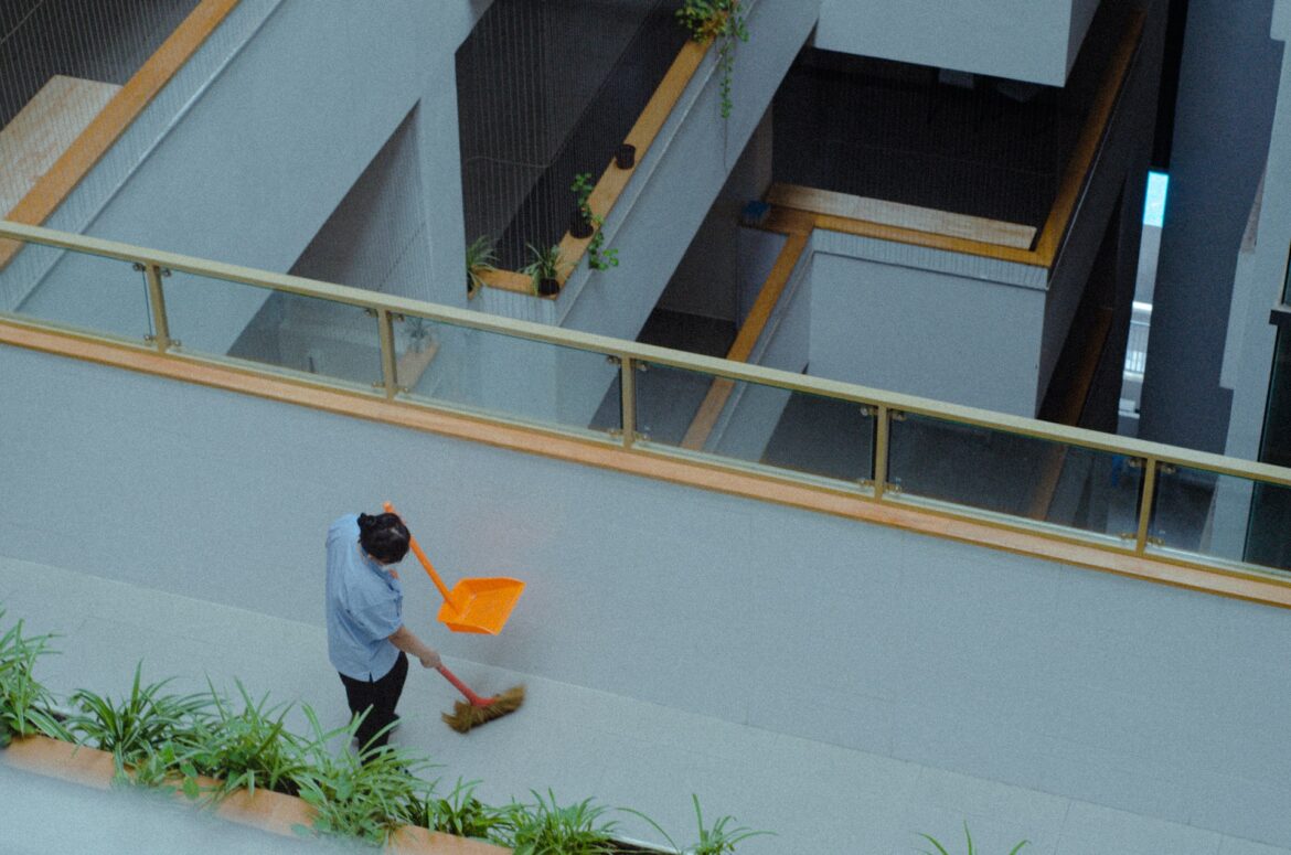 A person in a blue shirt sweeps a light gray floor with an orange dustpan in a modern multi-level atrium with plants and glass railings.] ,