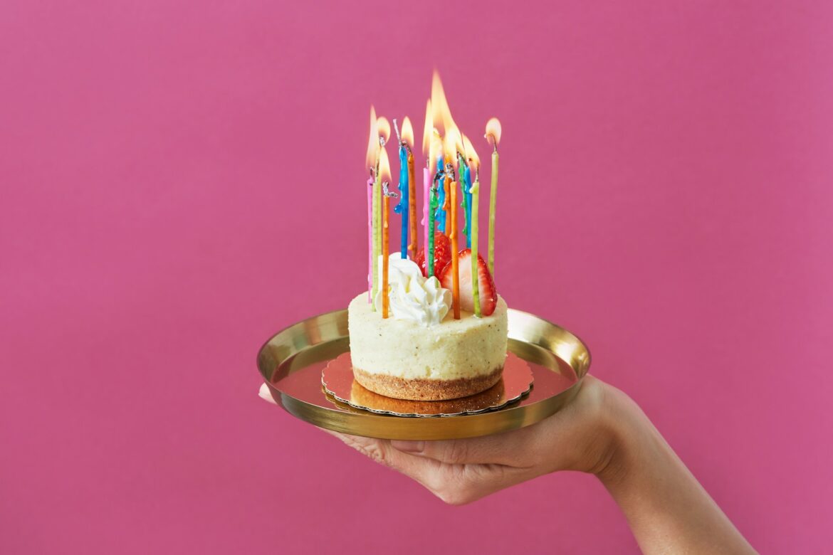 Birthday cake with many colorful lit candles on a gold plate against a pink background, held by a hand.