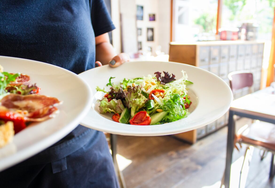 Server in a dark apron holds a plate of mixed salad in a bright cafe. (informative)