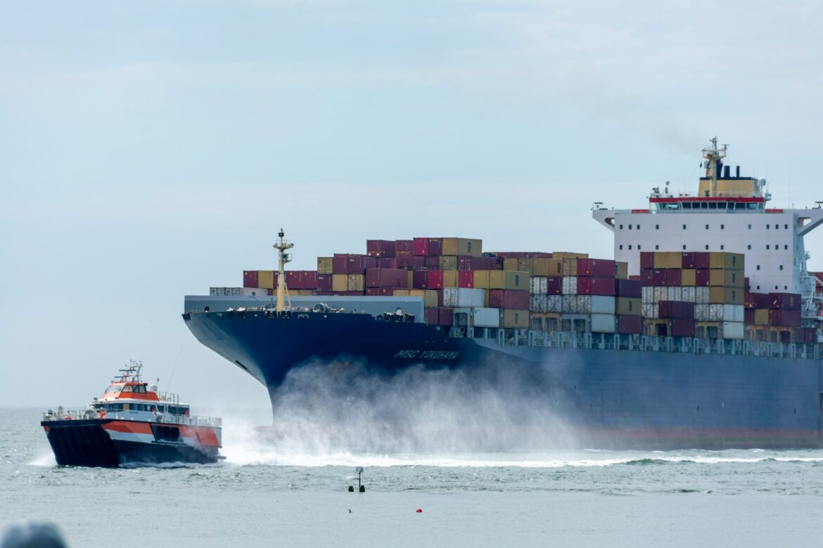 Large container ship with stacked colorful containers, guided by a small tugboat on choppy water under a clear sky.