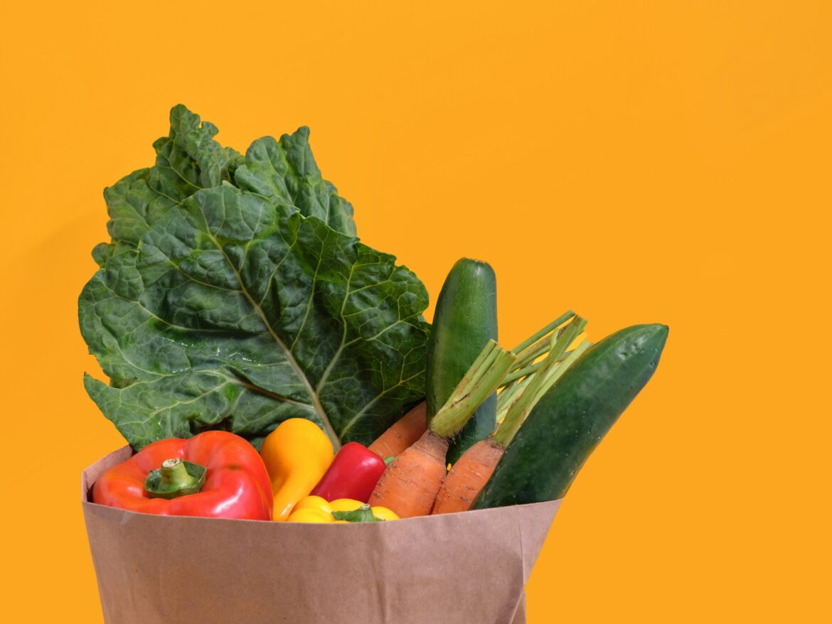 Paper bag filled with assorted fresh vegetables (peppers, cucumbers, carrots, leafy greens) against an orange background.