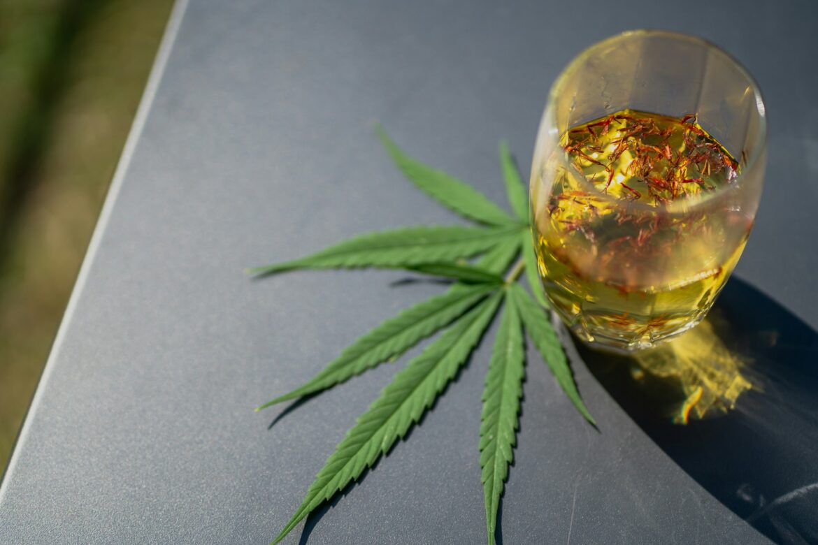 Glass of amber whiskey beside a cannabis leaf on a gray surface.