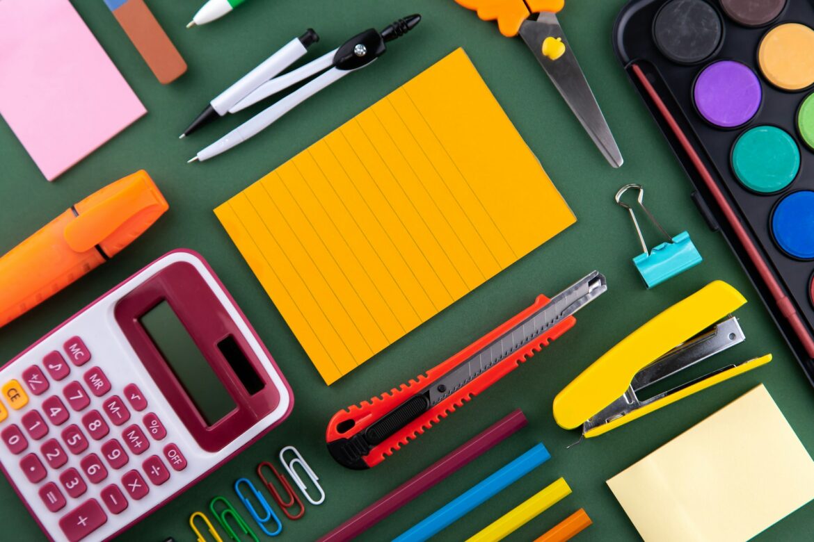 Colorful school supplies arranged diagonally: yellow notepad, pens, scissors, ruler, tape, binder clips, and a pink calculator on green background