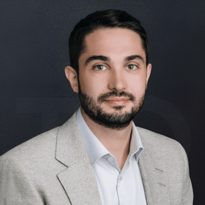 Portrait of a man with dark hair and a beard, wearing a light gray blazer and white shirt, against a dark textured background