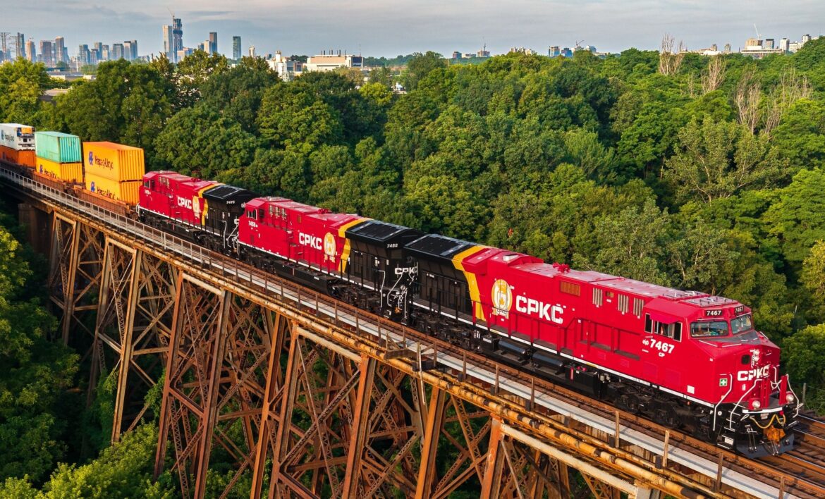 Red CPKC freight train with colorful containers crossing a tall steel trestle bridge over a dense green forest, city skyline in the distance.