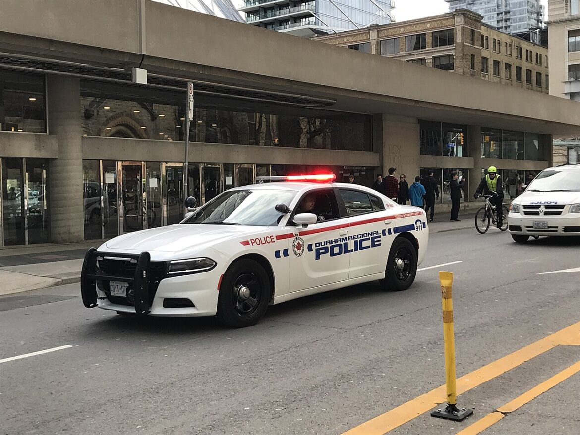 White Durham Regional Police car with red lights on, parked on a city street; officers and pedestrians nearby in front of a glass-fronted building.