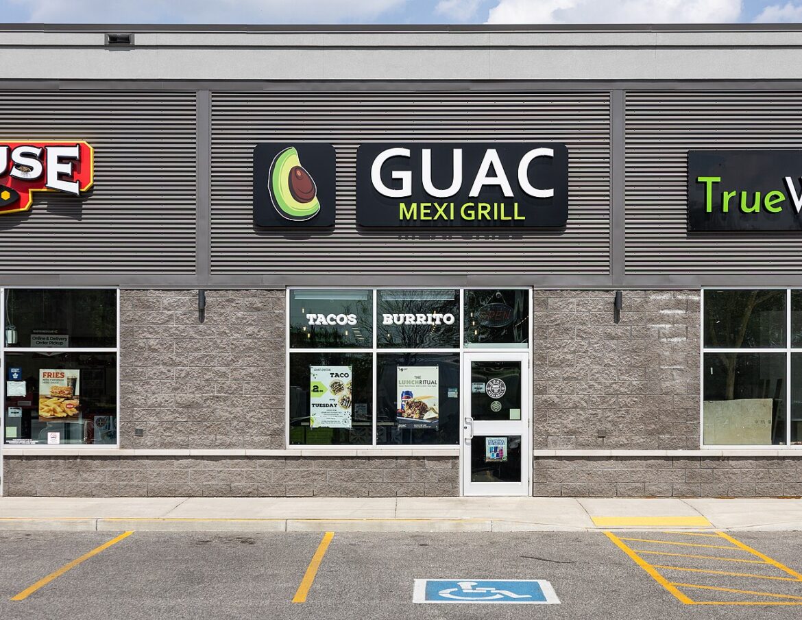 GUAC Mexi Grill storefront with avocado logo; windows show tacos and burrito posters, and a handicap parking space in the foreground.