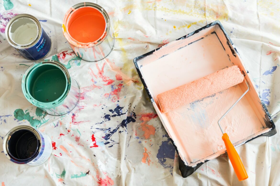 Overhead view of painting supplies on a paint-splattered drop cloth: open cans of orange, teal, and green paint and a pink paint tray with a roller.