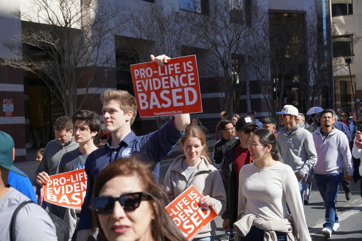 Crowd of protesters marching outdoors, holding orange signs that read 'PRO-LIFE IS EVIDENCE BASED.'