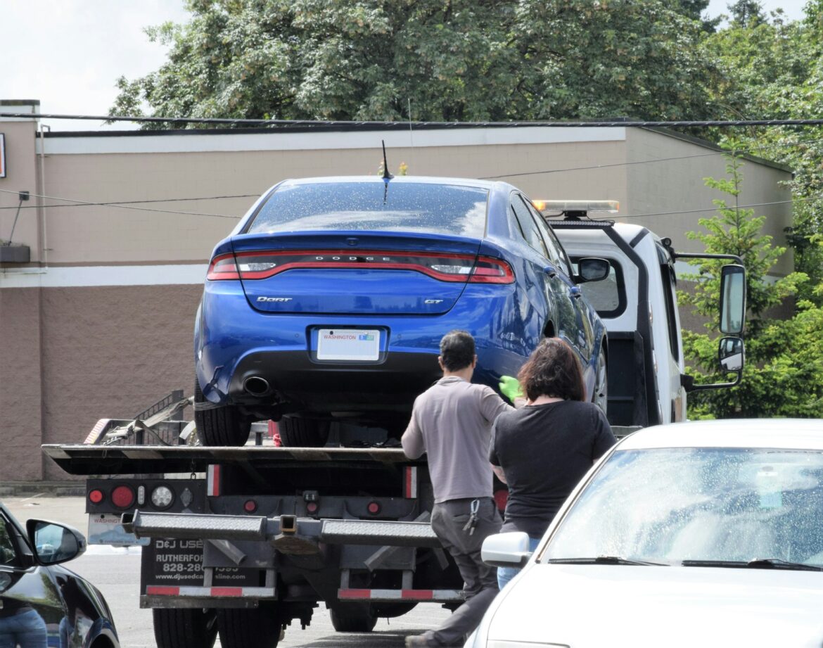 Blue Dodge Dart GT loaded onto a flatbed tow truck, with two people standing nearby in a parking lot.