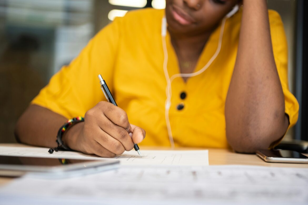 Person in a bright yellow shirt writes with a pen at a desk, papers spread out, wearing white earphones around the neck/face area.