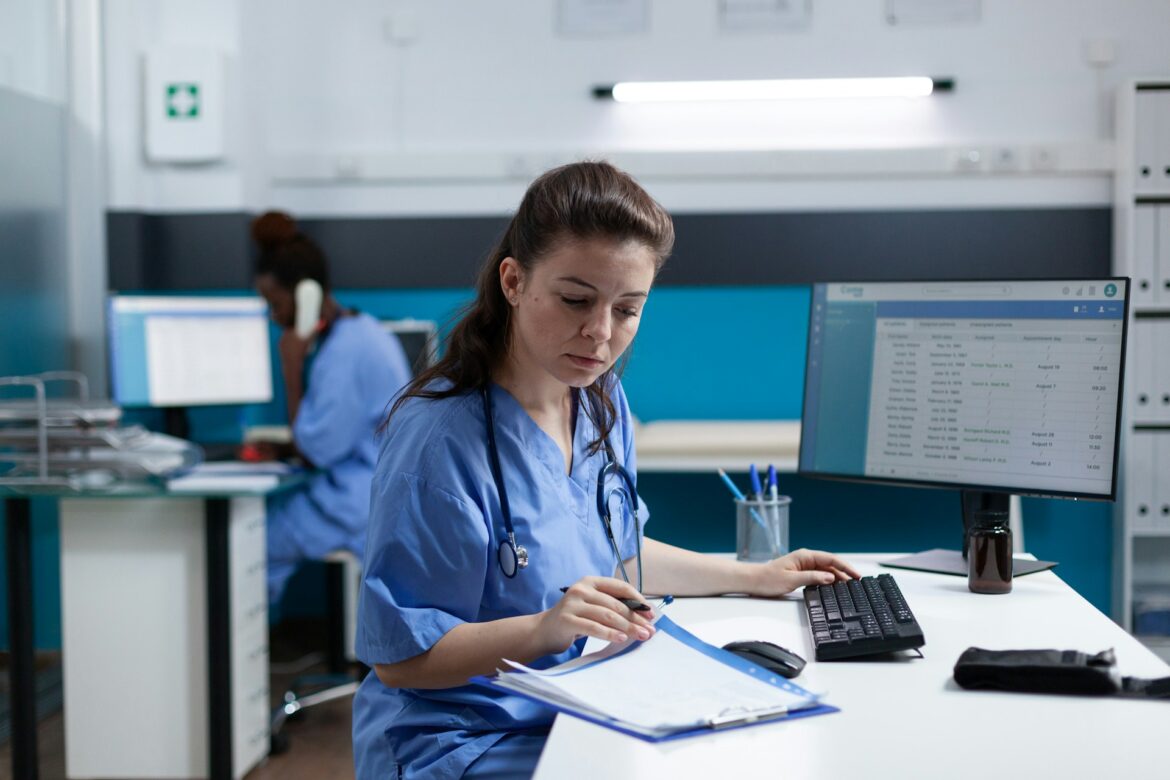 Nurse in blue scrubs works at a desk with a keyboard, papers, and a monitor showing a spreadsheet.