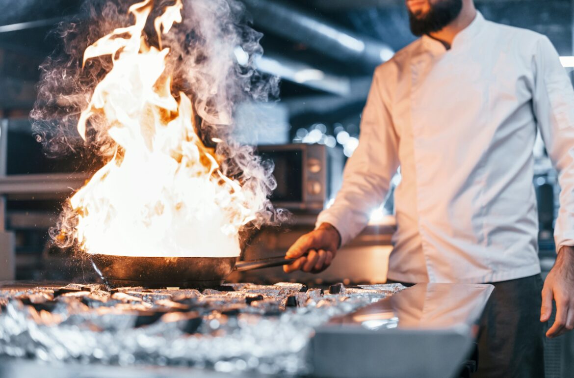 Chef in a white jacket cooking over a gas stove with large flames in a professional kitchen.