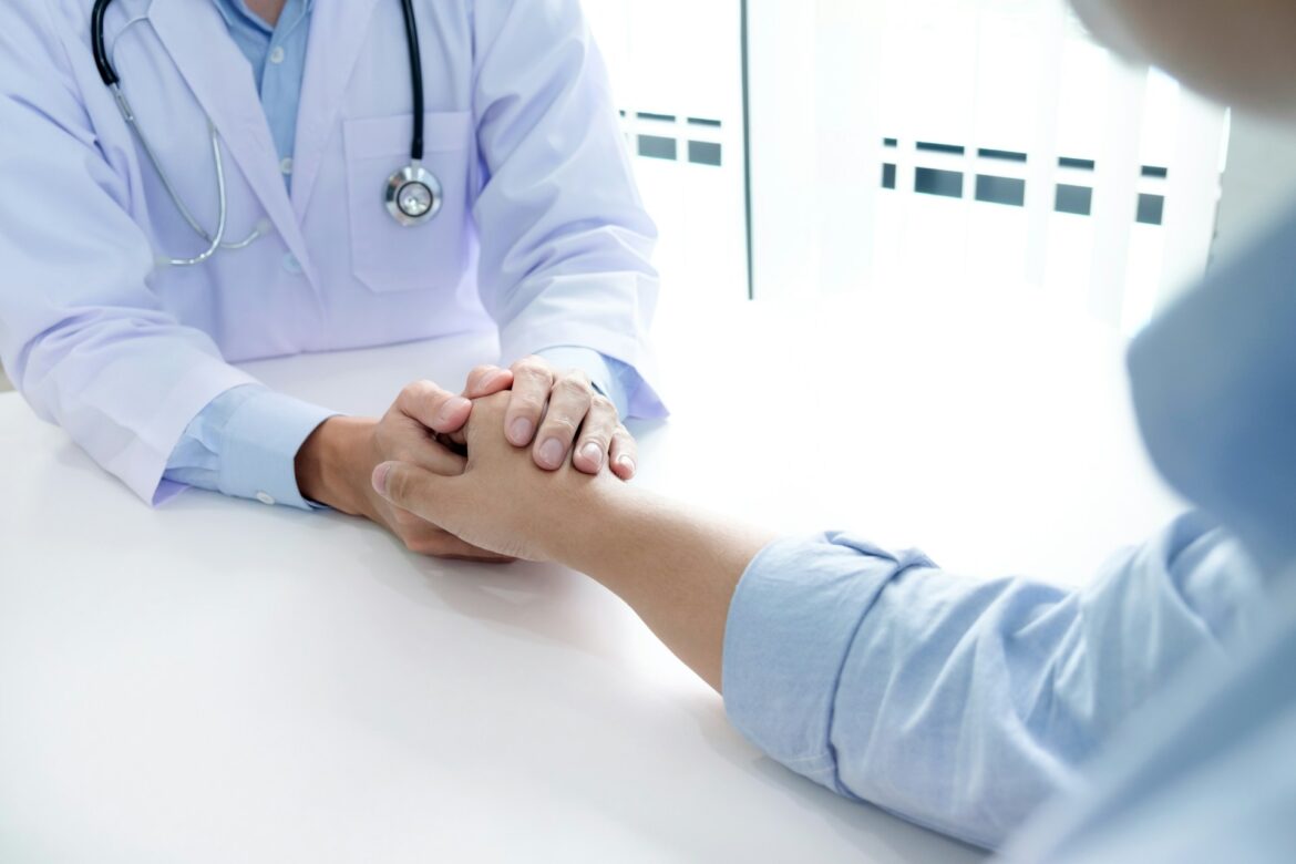 Doctor in a white coat with a stethoscope holds a patient’s hands across a clinic table, offering reassurance.