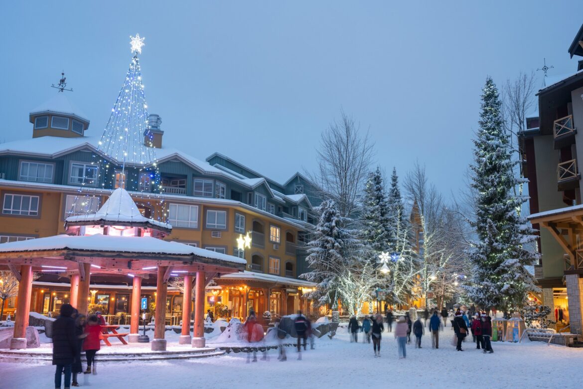 Snowy town square at dusk with a tall lit Christmas tree and people walking under festive lights.